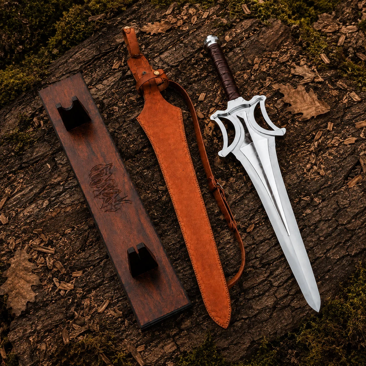 Top-down display of the He-Man sword, brown sheath and wooden plaque on bark with moss and dry leaves