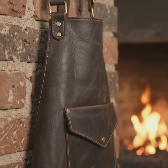 Anvil Arc Blacksmith Leather Apron — obsidian black cowhide, adjustable cross-back straps, antique brass-finish details and practical front pockets. Built around the traditional look of blacksmith workshop wear.