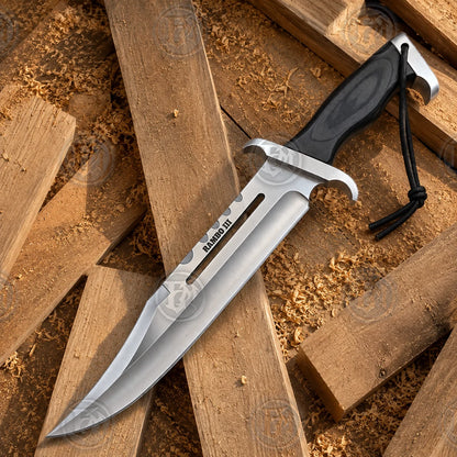 Full product view of the Rambo III survival Bowie knife replica on rustic wood, showing the curved blade profile and brownhandle.