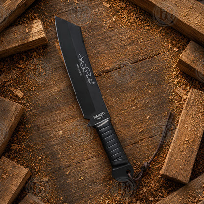 Full-length Rambo IV movie machete displayed on rustic timber with black blade, signature detailing and lanyard.